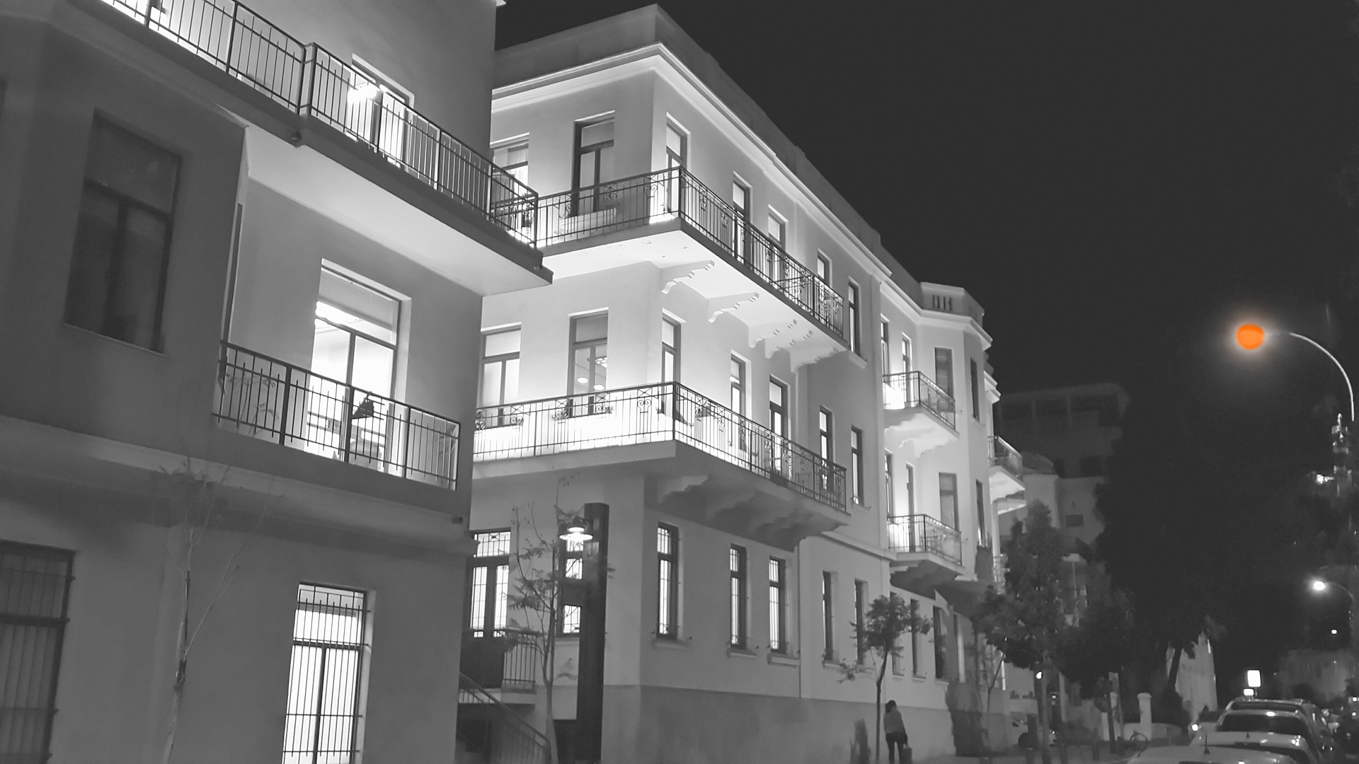 Tel Aviv Bauhaus street at night in black and white, one street lamp in orange. Photo: Юката, CC BY-SA 3.0, via Wikimedia Commons