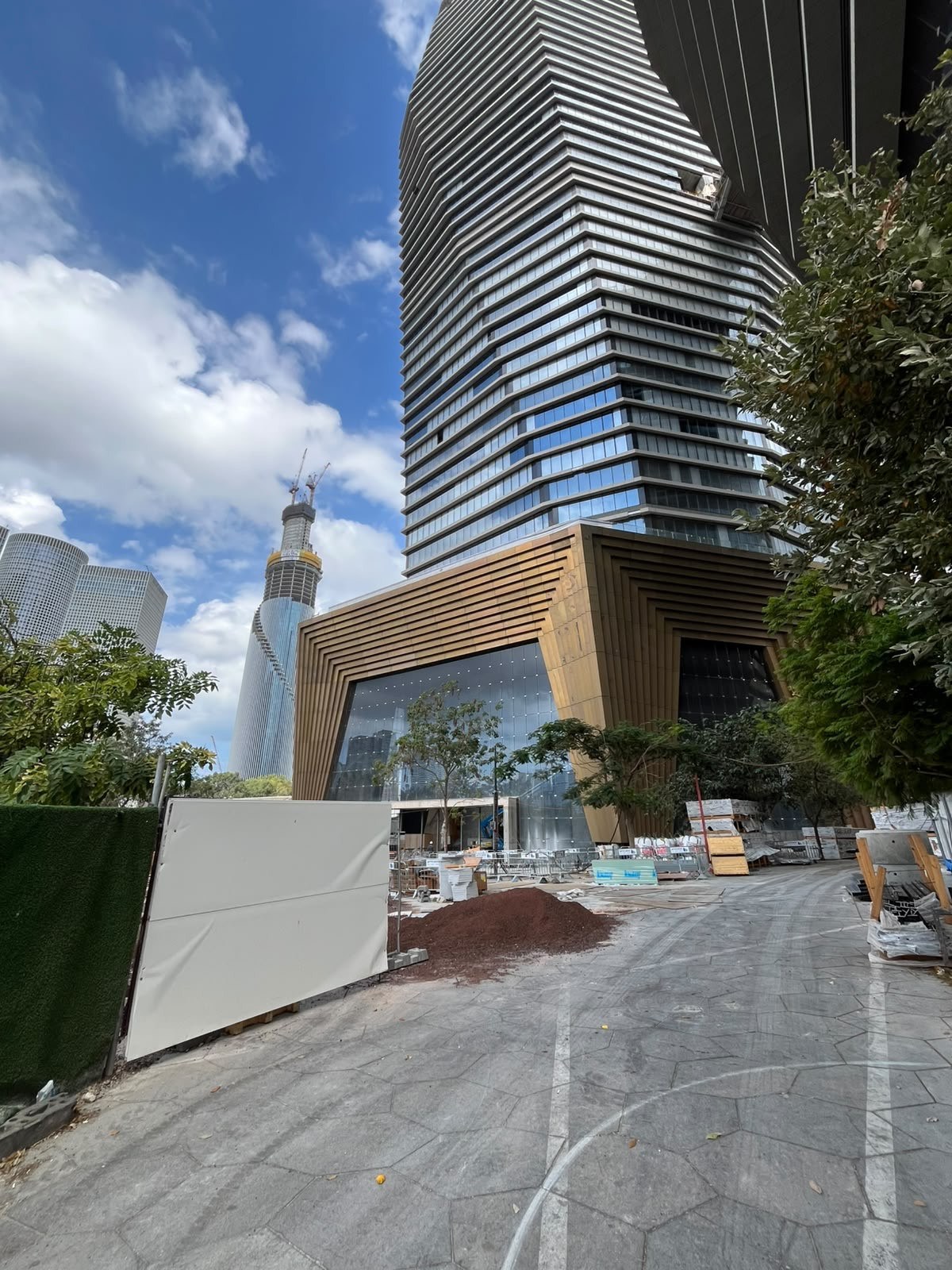 Looking up at a tower rising above a multi-layered bronze-toned entrance frame, with the Azrieli towers on the Tel Aviv skyline behind