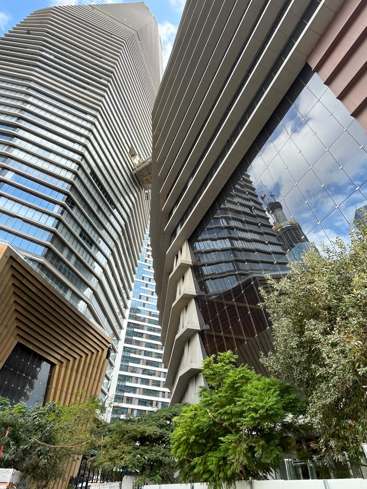 Looking up between two towers joined by a high skybridge, framed by trees and blue sky